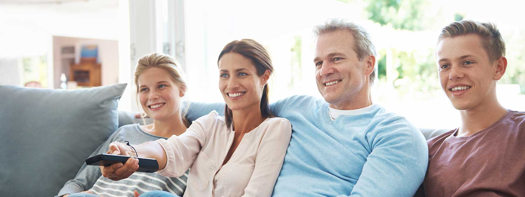 A family of four sits together on a couch, watching television and smiling, with one person holding a remote control.