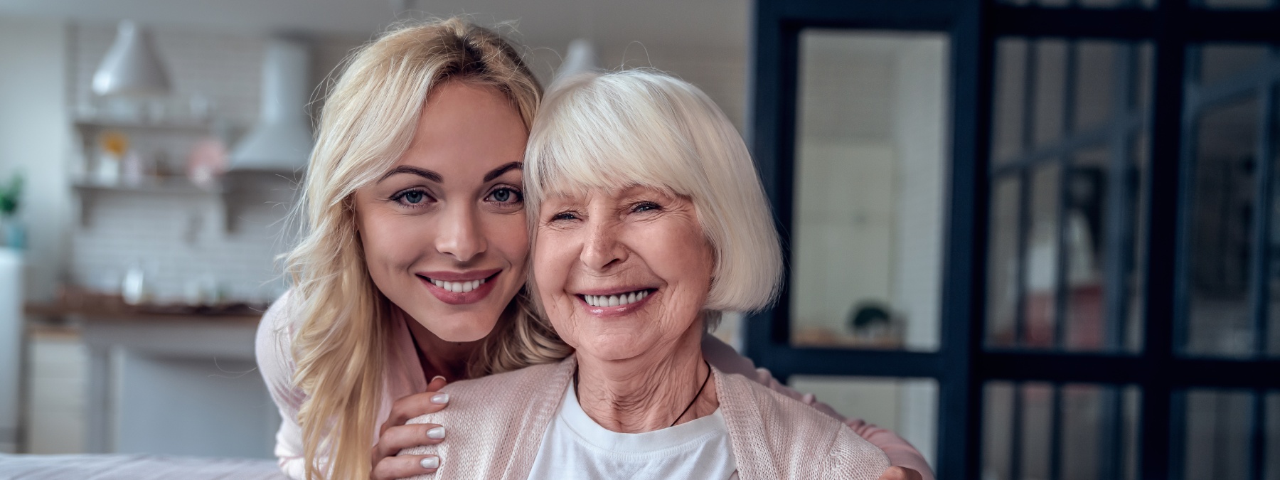 A younger woman and an older woman smile closely together, embracing with a cozy home setting in the background.